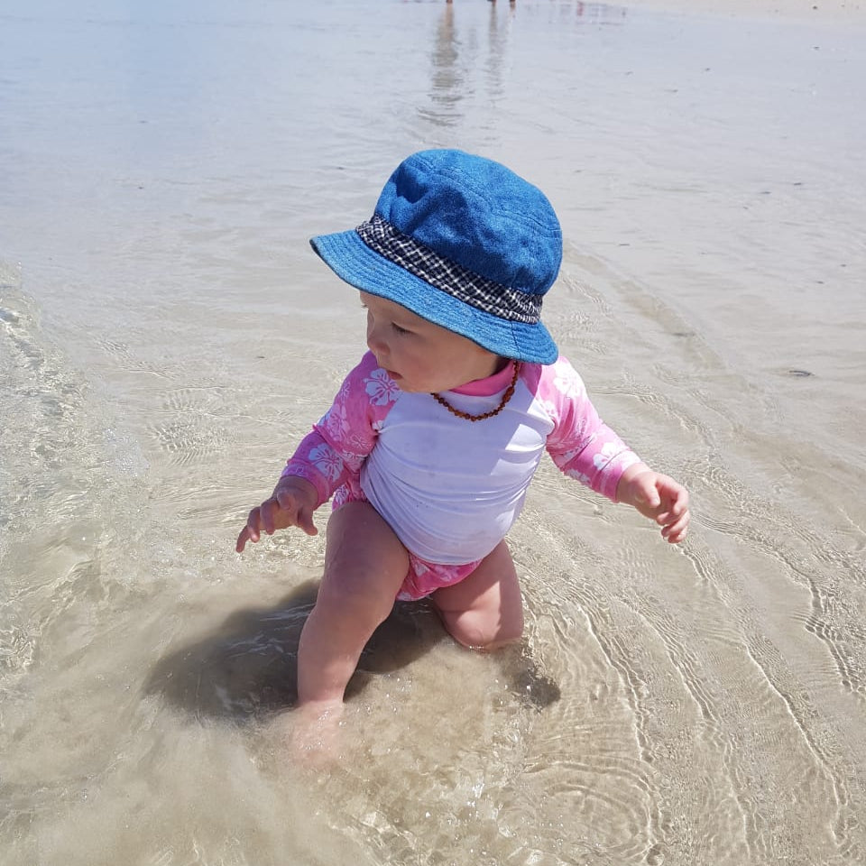 A baby wearing a blue sun hat and a pink and white long-sleeve swimsuit stands in shallow beach water on a sandy shore.