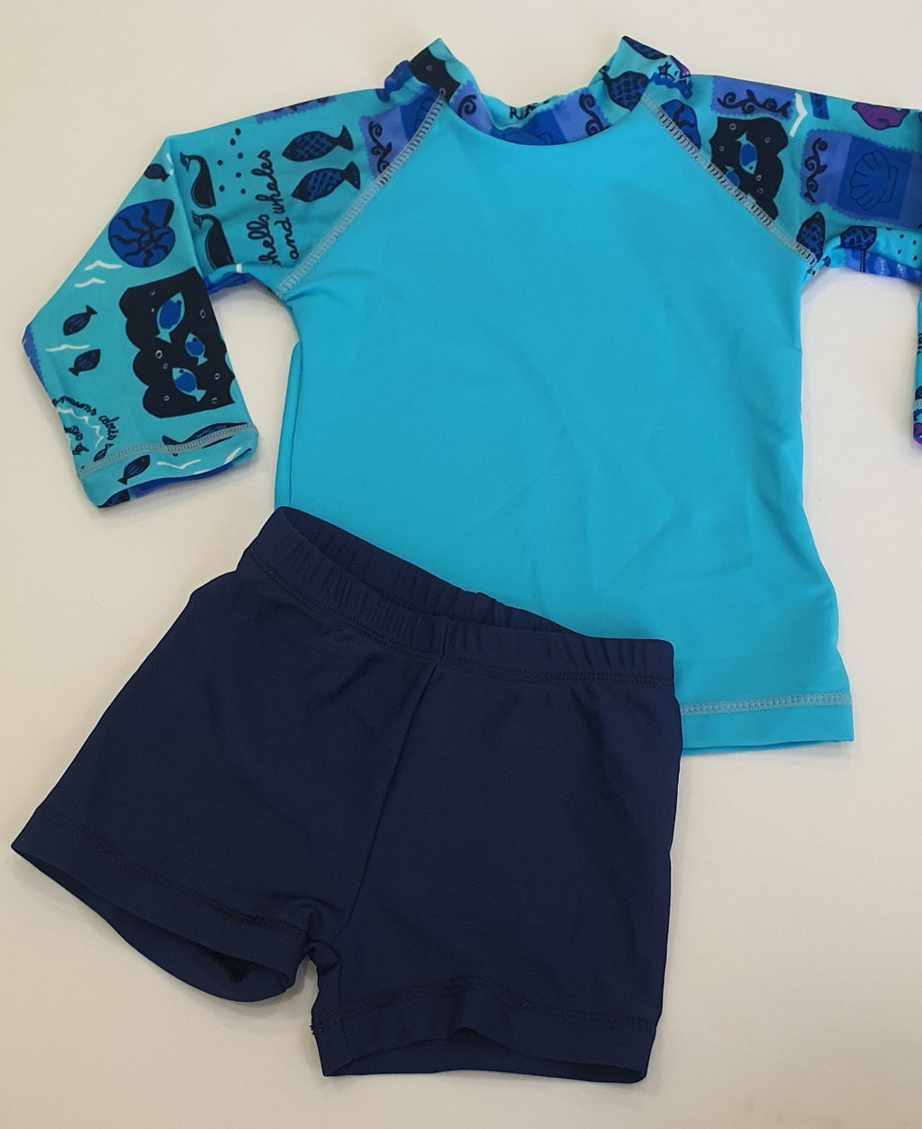 A children's two-piece swim outfit laid flat on a light beige background, featuring a bright turquoise long-sleeve rash guard with blue and black fish-patterned sleeves and navy blue swim shorts.