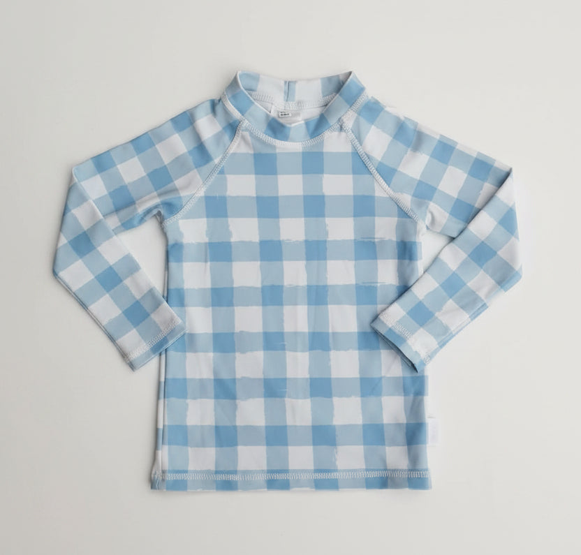 A long-sleeve children's rash guard or swim shirt with a light blue and white gingham check pattern, laid flat on a white textured fabric background.