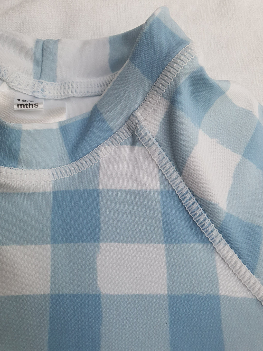 Close-up of a baby long-sleeve rashvest featuring a light blue and white gingham plaid pattern, with white ribbed trim and a visible 'to mths' brand label on the inner neck.