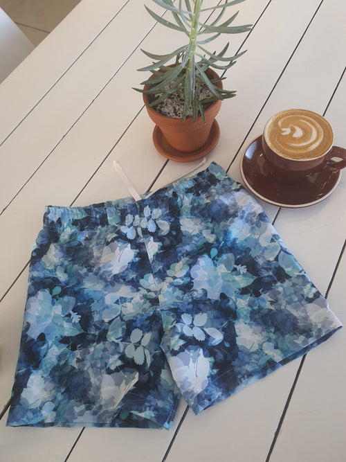 Blue floral swim shorts with built in nappy on a white wooden table with a plant and coffee cup.