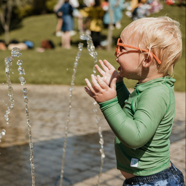 A young child with blonde hair and orange glasses wearing a green long-sleeve rash guard swimsuit, standing in a water fountain at an outdoor park.