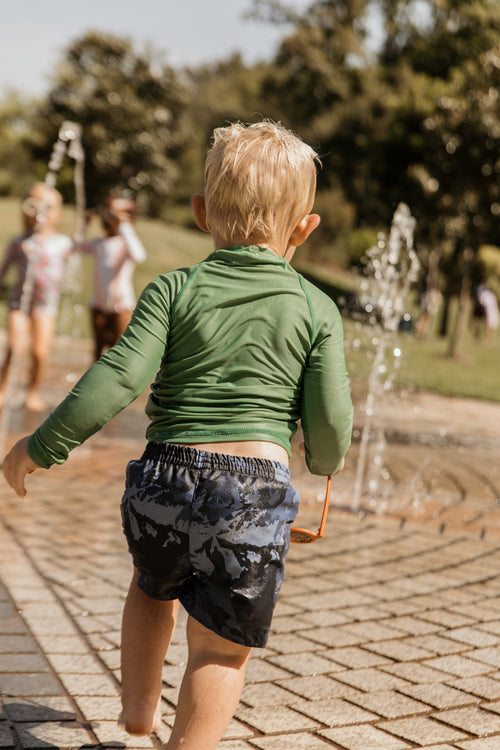 Child in green long-sleeve shirt and camouflage shorts  with built in swim nappy walking on a paved path with fountains and people in the background.