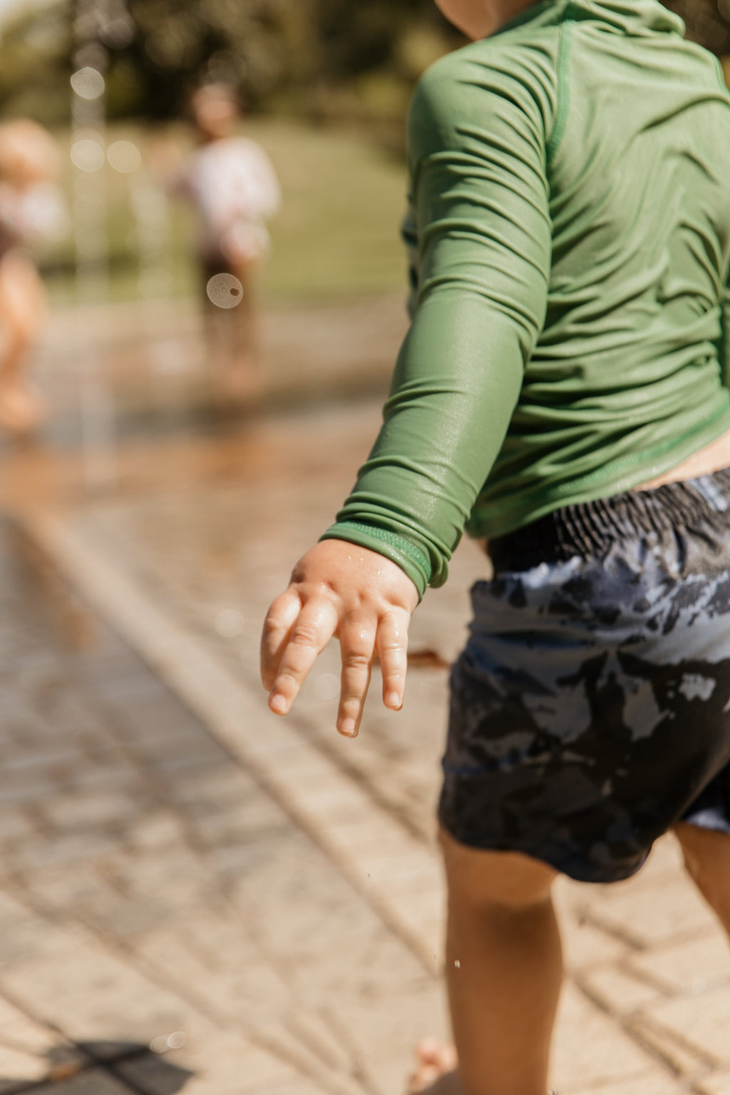 A child wearing a green long-sleeve rash guard and dark blue patterned swim trunks running through an outdoor water fountain splash pad.