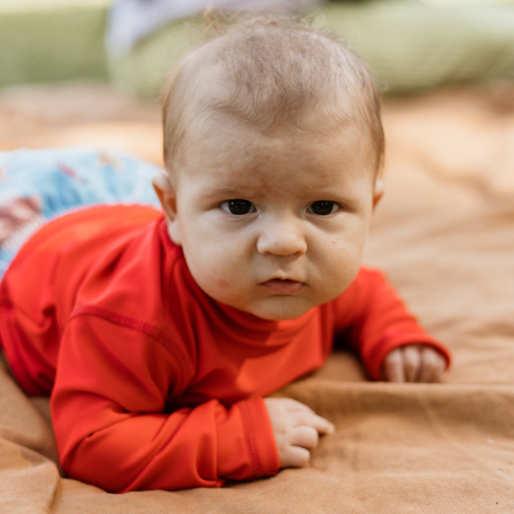 A baby lying on their stomach, wearing a bright red long-sleeve rash vest and light blue patterned pants, resting on a brown blanket.