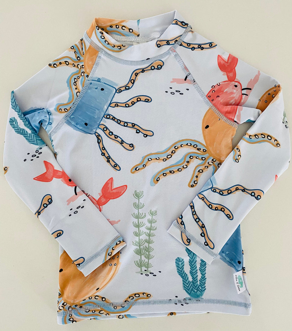 A long-sleeve children's swim shirt with an all-over ocean-themed print featuring octopus tentacles, crabs, sea plants, and fish on a white background, laid flat on a light beige surface.