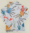 A long-sleeve children's swim shirt with an all-over ocean-themed print featuring octopus tentacles, crabs, sea plants, and fish on a white background, laid flat on a light beige surface.