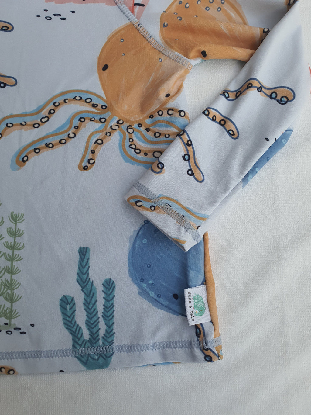A close-up of a children's long-sleeve top or pajama featuring a playful octopus and seaweed print on a white base. The design includes a large tan octopus with black and blue details, blue watercolor-style shapes, and green seaweed.