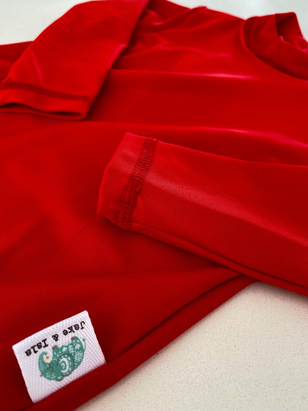 A close-up of a bright red long-sleeve red rash vest laid flat on a light beige surface, showing the sleeve cuff, fabric texture, and a small sewn-in label that reads 'Jube & Jada'.