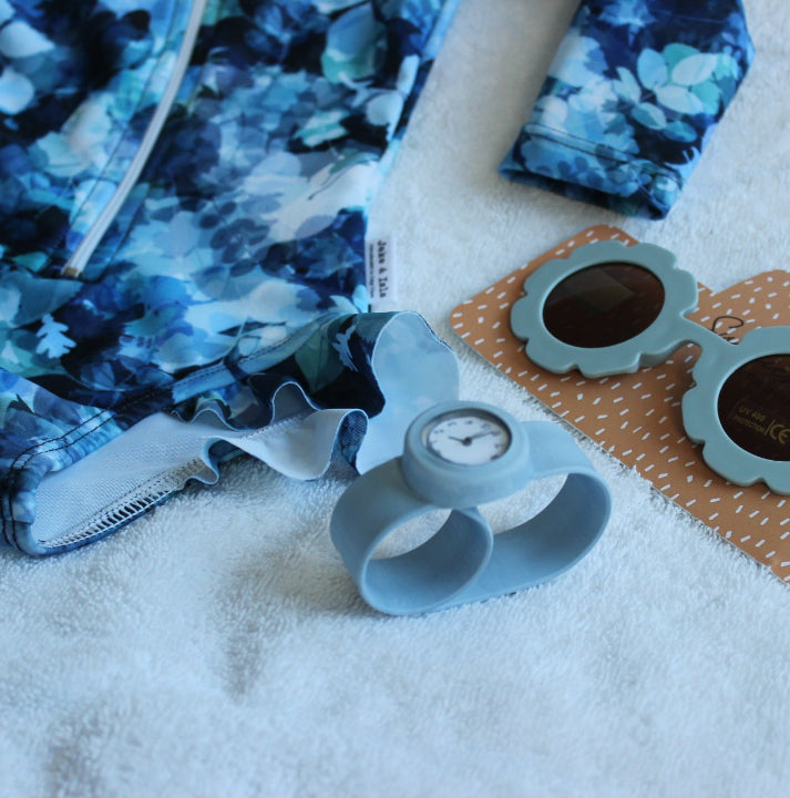 A flat-lay of a blue and white floral children's swimwear set alongside a light blue silicone wrist watch and flower-shaped sunglasses on a white textured towel.