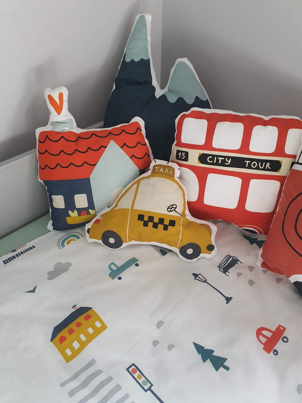 Set of colorful children's pillows shaped like a house, mountain, taxi, and city tour bus on a bed with a patterned sheet.