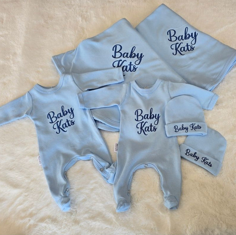 A baby clothing set laid flat on a cream faux-fur surface, including a light blue long-sleeve footless bodysuit, matching long-sleeve bodysuit, light blue bonnet, and light blue hat, all printed with 'Baby Kats' in dark blue script.