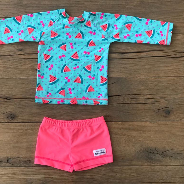A two-piece children's swim outfit laid flat on a wooden surface, featuring a turquoise long-sleeve top with a watermelon print and bright pink swim shorts.