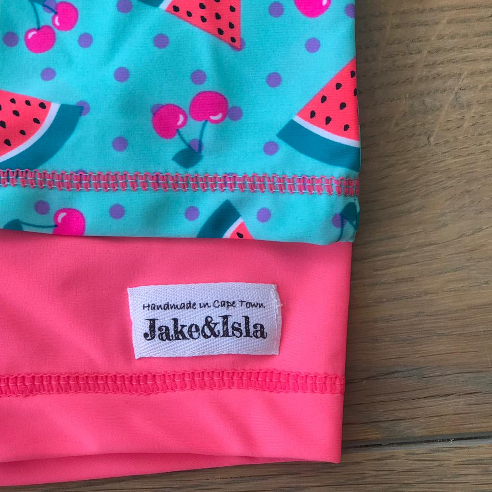 A folded fabric product, likely a swim cap or headband, featuring a bright pink solid section with a white 'Jake&Isla' brand label, and a turquoise section with a watermelon and cherry print, laid flat on a wooden surface.