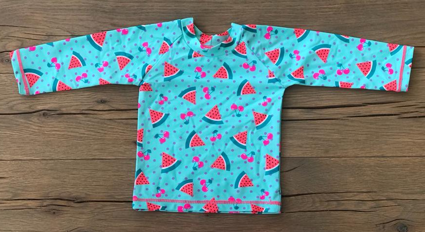 A long-sleeve children's top laid flat on a wooden surface, featuring a turquoise base with a repeating pattern of pink watermelon slices, red cherries, and small pink bows.