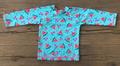 A long-sleeve children's top laid flat on a wooden surface, featuring a turquoise base with a repeating pattern of pink watermelon slices, red cherries, and small pink bows.