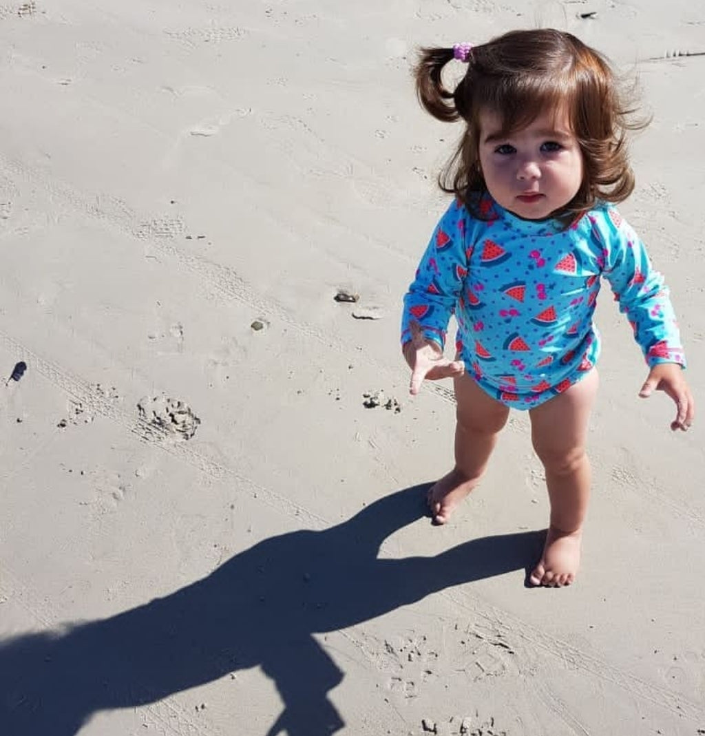 A toddler standing on a sandy beach wearing a light blue long-sleeve swim shirt with a red watermelon print. The child has brown hair in a small ponytail and is barefoot, with a long shadow cast on the sand.