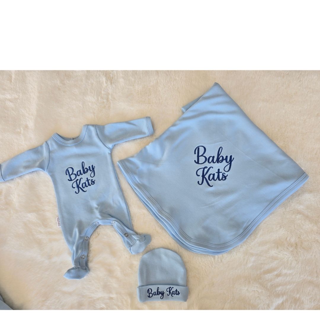 A baby gift set featuring a light blue footed sleeper, a matching square blanket, and a small matching beanie, all printed with the text 'Baby Kats' in navy blue script, laid out on a beige faux fur surface.