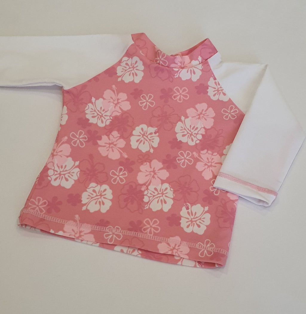 A pink sleeveless top with a white hawaiin floral print and long white sleeves, laid flat on a light beige background. jake and isla swimwear 