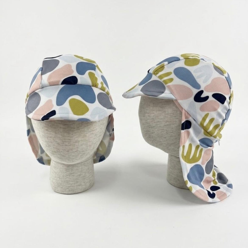 Two views of a children's sun hat with a neck flap, displayed on gray mannequin heads against a white background. The hat features an abstract organic pattern in blue, pink, olive green, and navy on a white base.