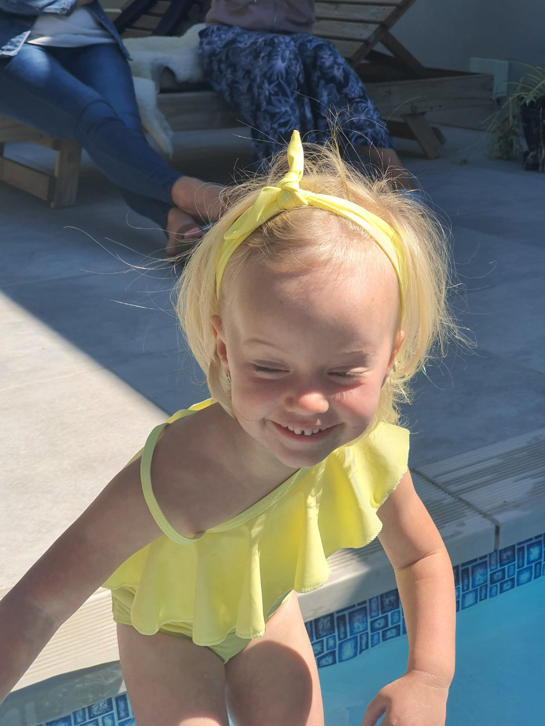 Child in a yellow swimsuit and headband standing by a pool
