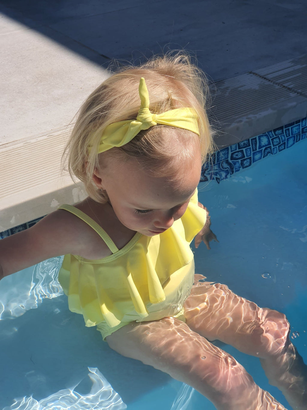 Child in a yellow swimsuit and headband standing by a pool